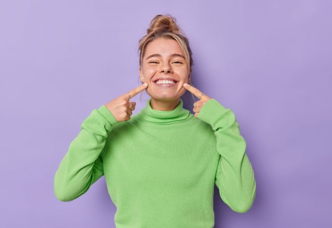 beautiful-happy-fair-haired-european-woman-points-index-fingers-mouth-forces-cheerful-smile-shows-perfect-white-teeth-wears-green-jumper-being-good-mood-isolated-purple-background-min